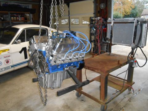 1972 Ford Bronco - A V8 engine with blue spark plug wires is suspended by chains next to a classic white Ford Mustang GT 350. The engine is mounted on a rolli…