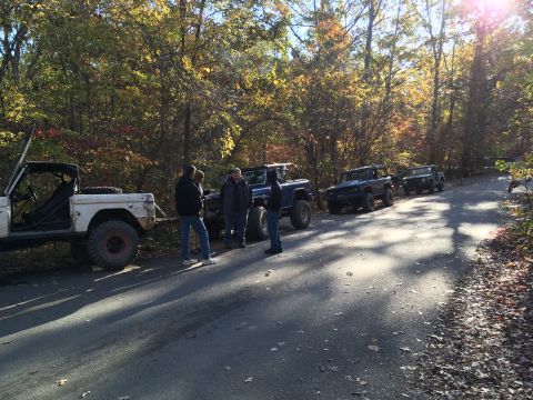 1979 Ford Bronco - A group of people stand around several off-road vehicles parked on a paved road in a wooded area during autumn. The vehicles appear to be F…