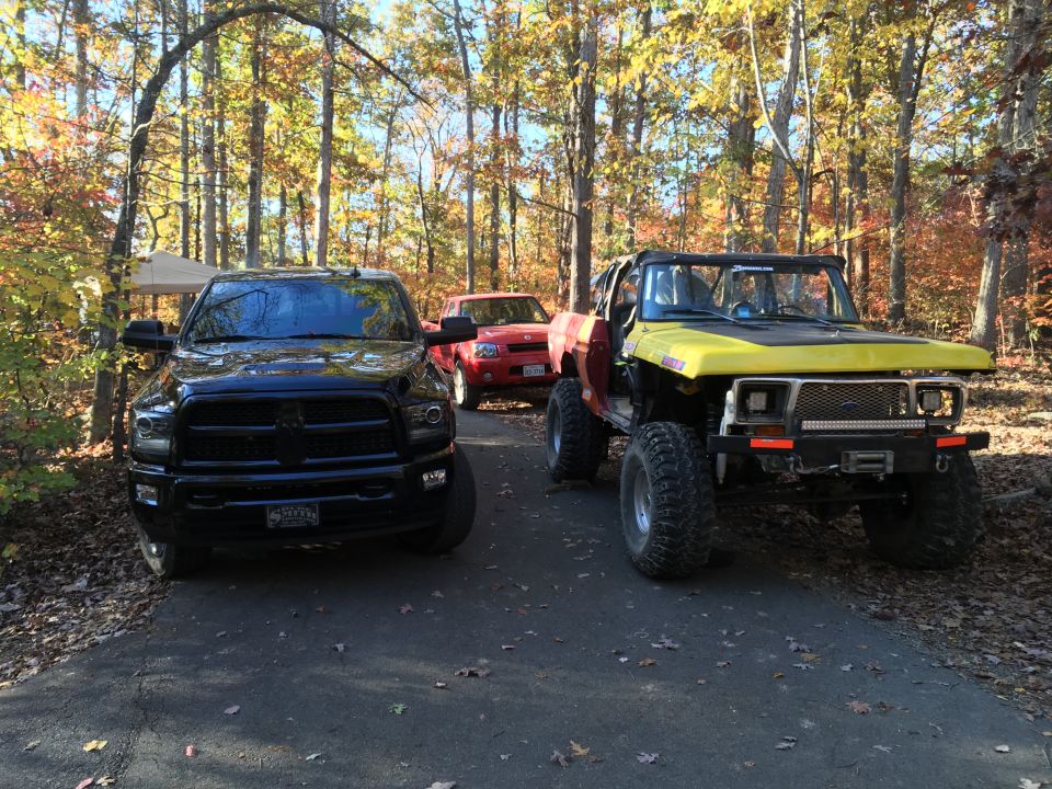 Black Ram truck, red pickup, and yellow/red custom Ford Bronco in a wooded area in fall.