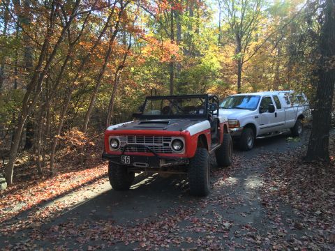 1979 Ford Bronco - A red and white Ford Bronco is parked on a leaf-covered dirt road next to a white Ford pickup truck with a camper shell. The Bronco has a b…