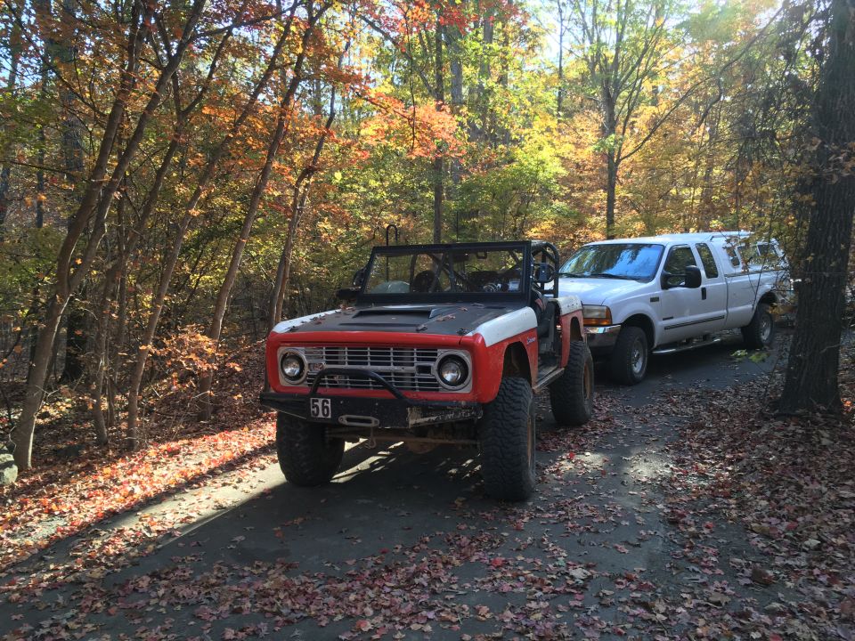 Red and white Ford Bronco parked on a dirt road with fall leaves, next to a white Ford pickup truck.
