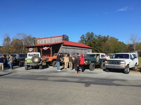 1979 Ford Bronco - A group of people stand around several vintage Ford Broncos and other off-road vehicles parked in front of a wooden building with a red roo…