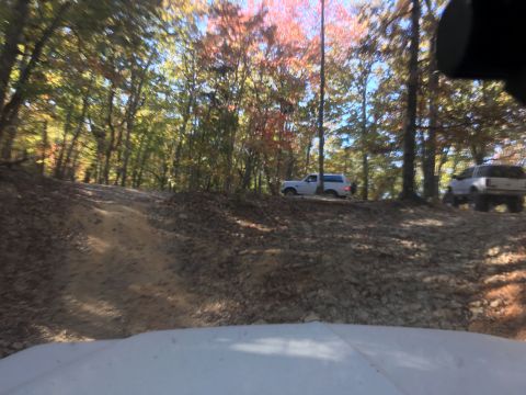 1979 Ford Bronco - Two white SUVs are driving on a dirt road through a forest with autumn foliage. The vehicle in the foreground is partially visible from the…