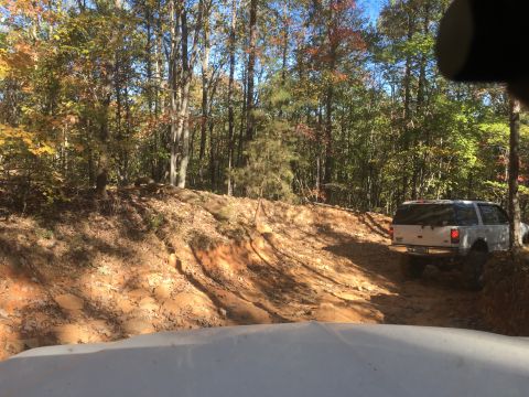 1979 Ford Bronco - A white SUV is driving up a steep, rutted dirt trail in a wooded area during autumn. The vehicle is viewed from behind and slightly to the…