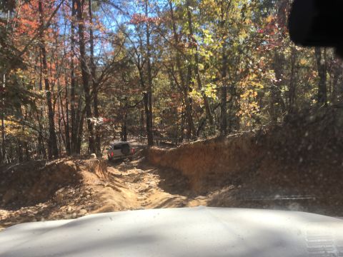 1979 Ford Bronco - A white SUV is driving up a rutted, dirt trail through an autumn forest. The vehicle is viewed from behind and slightly to the side, naviga…