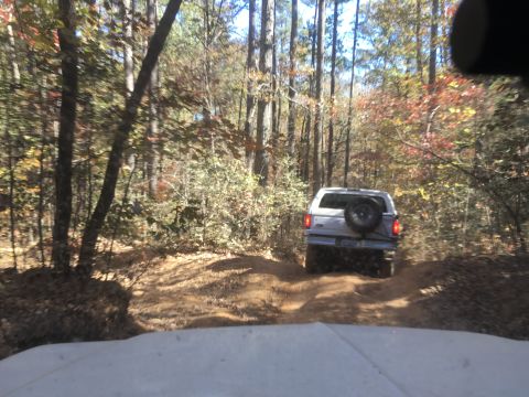 1979 Ford Bronco - A silver SUV drives down a dirt path in a wooded area during the fall. The vehicle has a spare tire mounted on the rear.