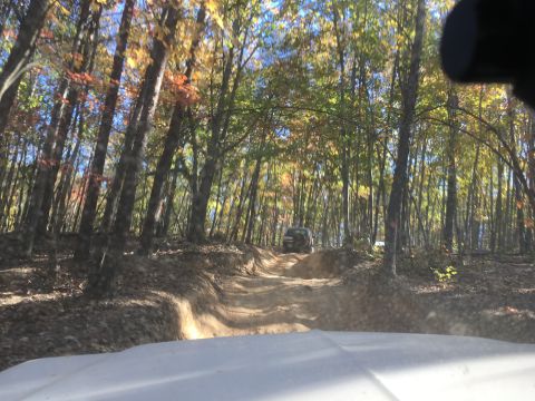 1979 Ford Bronco - A dark colored SUV drives up a dirt trail through a forest with autumn foliage. The vehicle is seen from the perspective of another vehicle…