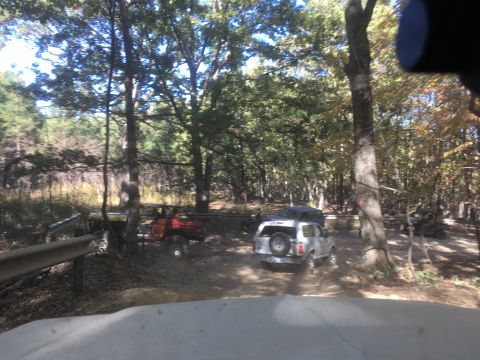 1979 Ford Bronco - A white Ford Bronco with a spare tire on the back is parked on a dirt trail in a wooded area. A red off-road vehicle is parked behind it.