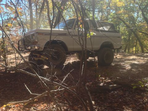 1979 Ford Bronco - A white Ford Bronco with a soft top is navigating a dirt path through a wooded area during autumn. The vehicle has a brush guard on the fro…