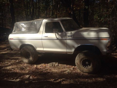 1979 Ford Bronco - A white 1970s Ford Bronco with a soft top is parked on a dirt path in a wooded area. The vehicle has large off-road tires and is angled to…