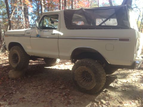1979 Ford Bronco - A white 1970s Ford Bronco with a soft top is parked off-road on a dirt path surrounded by trees with autumn foliage. The front passenger si…