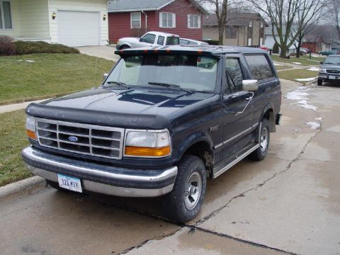 1995 Ford Bronco - A dark blue Ford Bronco is parked on a cracked asphalt driveway in front of a house.