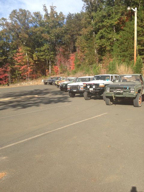 1979 Ford Bronco - A line of off-road modified SUVs are parked in a lot with trees showing fall colors in the background. The closest vehicle is a dark green…