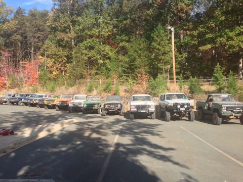 1979 Ford Bronco - A line of off-road modified SUVs and trucks are parked in a lot with a wooded area in the background. The vehicles are mostly older models…