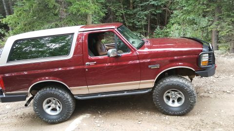 1996 Ford Bronco - A red 1996 Ford Bronco with a tan lower body accent is parked on a dirt road in a wooded area. The SUV has large off-road tires and a brush…