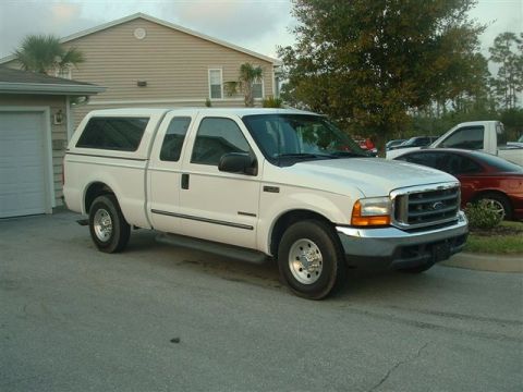 1986 Ford Bronco - A white pickup truck with a camper shell is parked in a driveway next to a house.