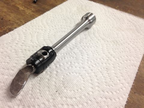 1979 Ford Bronco - A close-up of a custom tool with a metallic shaft and a black housing, featuring a curved metal blade at one end. The tool rests on a white…