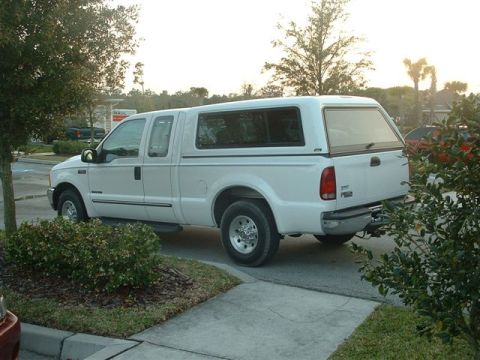 1986 Ford Bronco - A white pickup truck with a camper shell is parked on a paved surface next to a sidewalk and some bushes.