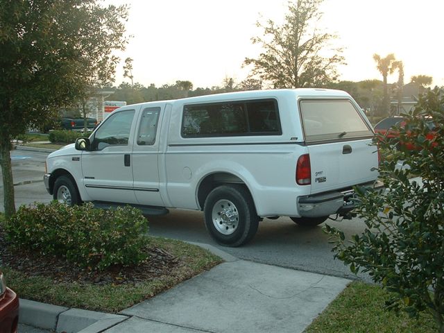 White pickup truck with a camper shell, seen from the side at a slight rear angle. The truck has four doors and appears to be in good condition