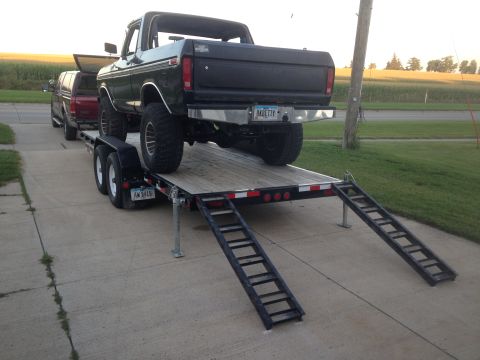 1979 Ford Bronco - A black 1979 Ford Bronco is loaded onto a flatbed trailer, parked on a concrete driveway next to a red pickup truck. The Bronco has large o…