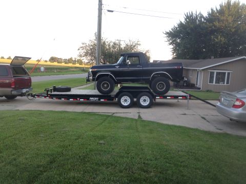 1979 Ford Bronco - A black Ford Bronco is loaded onto a tandem-axle car hauler trailer, which is being towed by a red SUV. A silver sedan is parked to the rig…
