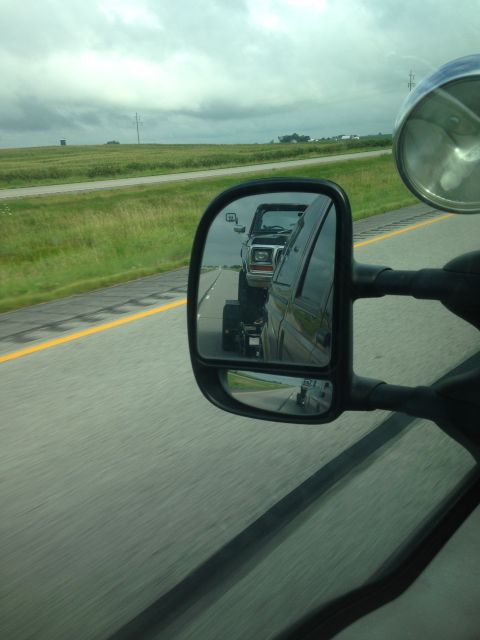 1979 Ford Bronco - The side mirror of a vehicle reflects a black Ford Bronco driving on a highway. The Bronco is towing a trailer.