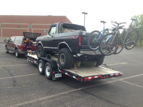 1979 Ford Bronco - A black Ford Bronco is loaded onto a flatbed trailer, with two bicycles mounted on the rear of the Bronco. A red Ford pickup truck is parke…