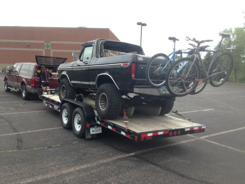 1979 Ford Bronco - A black Ford Bronco is loaded onto a flatbed trailer, with two bicycles mounted on a rack attached to the rear of the Bronco. A red Ford pi…