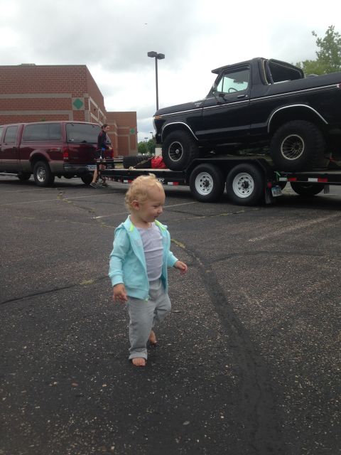 1979 Ford Bronco - A toddler walks barefoot on asphalt in front of a black Ford Bronco on a trailer. A red pickup truck is parked to the left.
