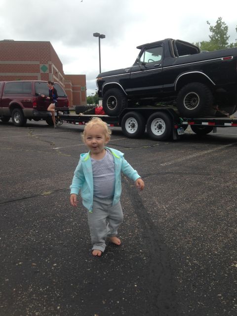 1979 Ford Bronco - A toddler stands barefoot on asphalt in front of a black Ford Bronco on a trailer. A red SUV is parked to the left of the trailer.
