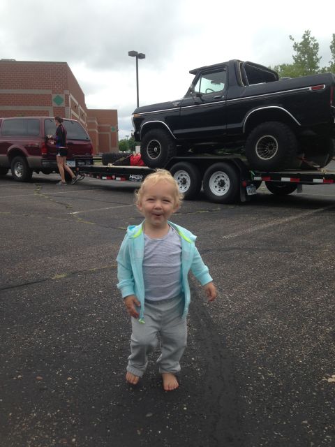 1979 Ford Bronco - A young child stands barefoot on asphalt in front of a black Ford Bronco on a trailer. A maroon SUV is parked to the left of the trailer.