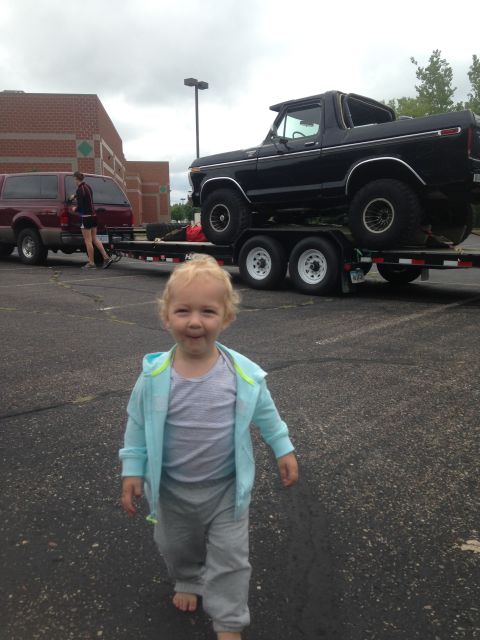1979 Ford Bronco - A young child walks towards the camera in a parking lot, with a black Ford Bronco on a trailer in the background.