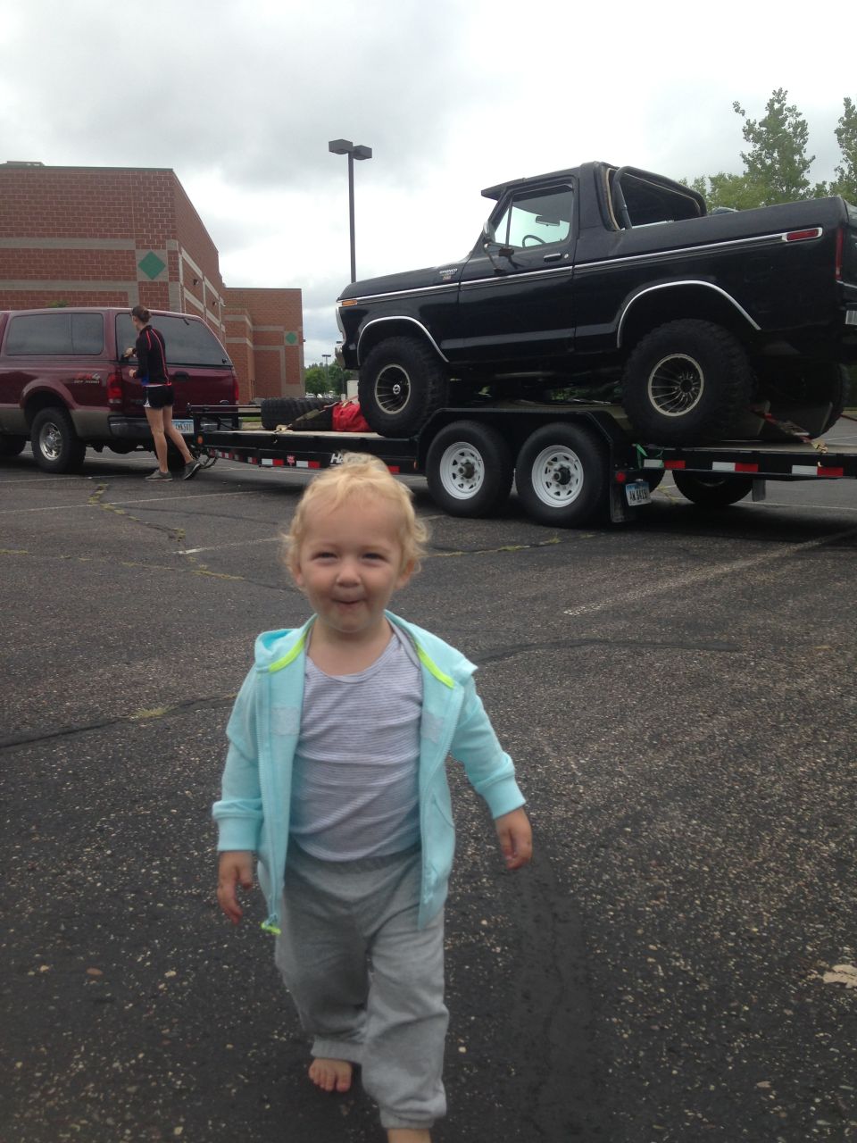 A toddler walks forward, a black Ford Bronco on a trailer is visible behind them.