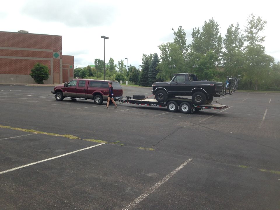 Black Ford Bronco on trailer towed by maroon Ford pickup truck, with bicycles on rear rack.