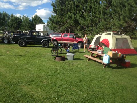 1979 Ford Bronco - A campsite scene features a black 1979 Ford Bronco parked on grass next to a red Ford pickup truck and a large tent. A picnic table with su…