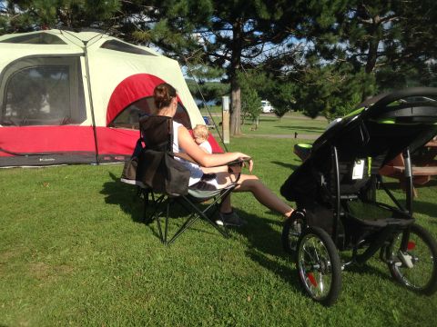 1979 Ford Bronco - A woman sits in a camping chair next to a tent with a baby in her lap. A stroller is parked next to her, and trees and a lake are visible i…