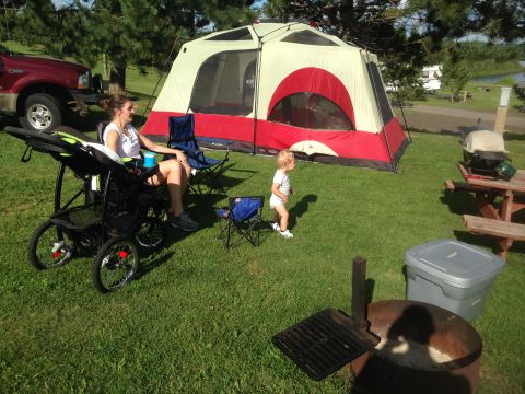 1979 Ford Bronco - A red Ford pickup truck is parked next to a large tent at a campsite. A woman is sitting in a camping chair next to a stroller, and a toddl…