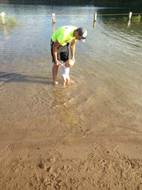 1979 Ford Bronco - A man in a neon green shirt and black shorts holds a baby's hands as they stand in shallow water near a sandy shore.