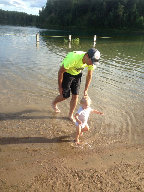1979 Ford Bronco - A man in a neon green shirt and black shorts bends down to help a toddler walk in shallow water at the edge of a lake.