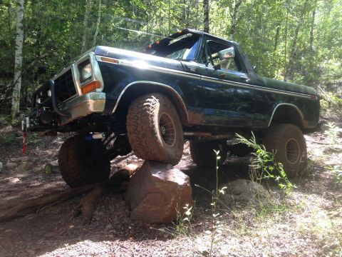 1979 Ford Bronco - A dark green 1979 Ford Bronco is navigating an off-road trail, with its front right tire perched on a large rock. The vehicle features a li…