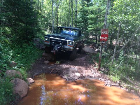 1979 Ford Bronco - A dark colored 1979 Ford Bronco is navigating a muddy, rocky trail through a forest. The vehicle is positioned at an angle, with its front…