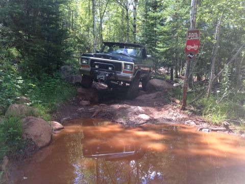 1979 Ford Bronco - A dark-colored Ford Bronco is navigating a muddy, rocky trail, with its front tires on large rocks and its front bumper close to a large mu…