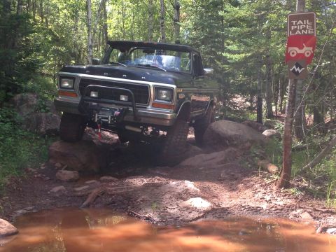 1979 Ford Bronco - A black Ford Bronco is navigating a muddy, wooded trail with large rocks and a puddle in the foreground. A sign indicating "Tail Pipe Lane"…