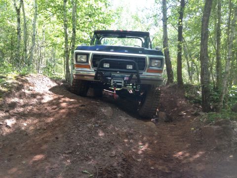 1979 Ford Bronco - A dark colored Ford Bronco is shown from the front, driving on a muddy, wooded trail. The vehicle has a brush guard with a winch mounted on…