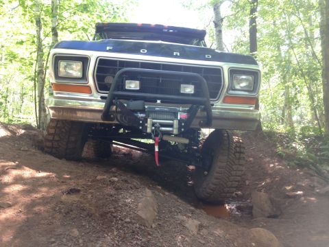 1979 Ford Bronco - A dark blue Ford Bronco is shown from the front, angled upwards on a dirt trail in a wooded area. The vehicle features a large winch mounte…