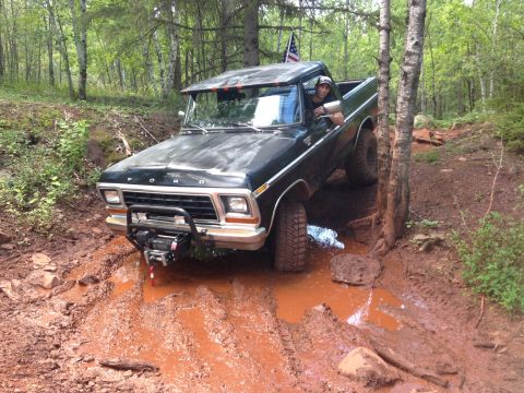 1979 Ford Bronco - A black Ford pickup truck with a winch mounted on the front bumper navigates a deep mud pit on a wooded trail. The driver is visible behind…