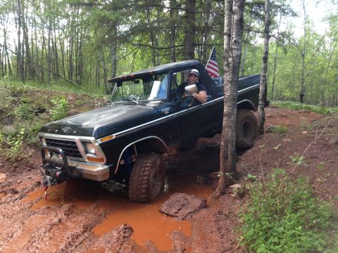 1979 Ford Bronco - A black 1979 Ford Bronco is navigating a muddy trail in a wooded area. The vehicle has an American flag displayed in the rear window and a…