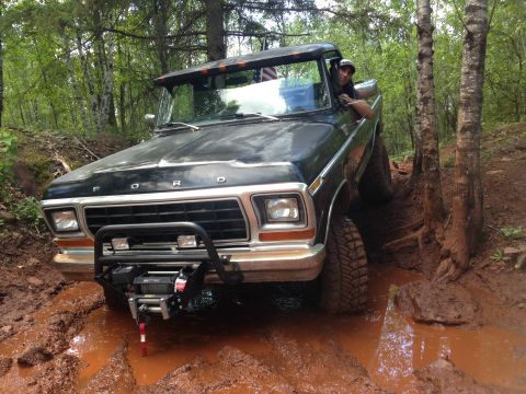 1979 Ford Bronco - A black 1970s Ford pickup truck is navigating a muddy trail in a forest, with its front tires deep in the mud and a winch mounted on the fr…