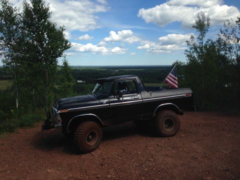 1979 Ford Bronco - A black 1979 Ford Bronco is parked on a dirt road with an American flag in the truck bed. The vehicle is viewed from the side at a 3/4 fron…