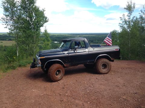 1979 Ford Bronco - A black 1979 Ford Bronco is parked on a dirt road overlooking a wooded landscape. An American flag is mounted on the tailgate of the vehicl…